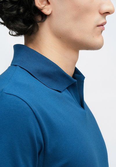Zils polo krekls ar ribotu apkakli un vieglu V veida izgriezumu, izgatavots no gludas auduma, demonstrējot pieguļošu siluetu.