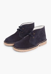 Botas de ante azul marino hasta el tobillo con suelas de goma marrón, que presentan cordones redondos y ojales dorados, textura suave y acentos de diseño mínimos.