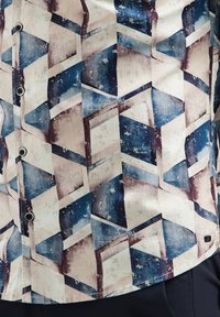 Geometrisch patroon overhemd met blauwe, beige en bruine tinten, voorzien van kubussen en schuine lijnen, met knopen, gedragen over donkere broek.