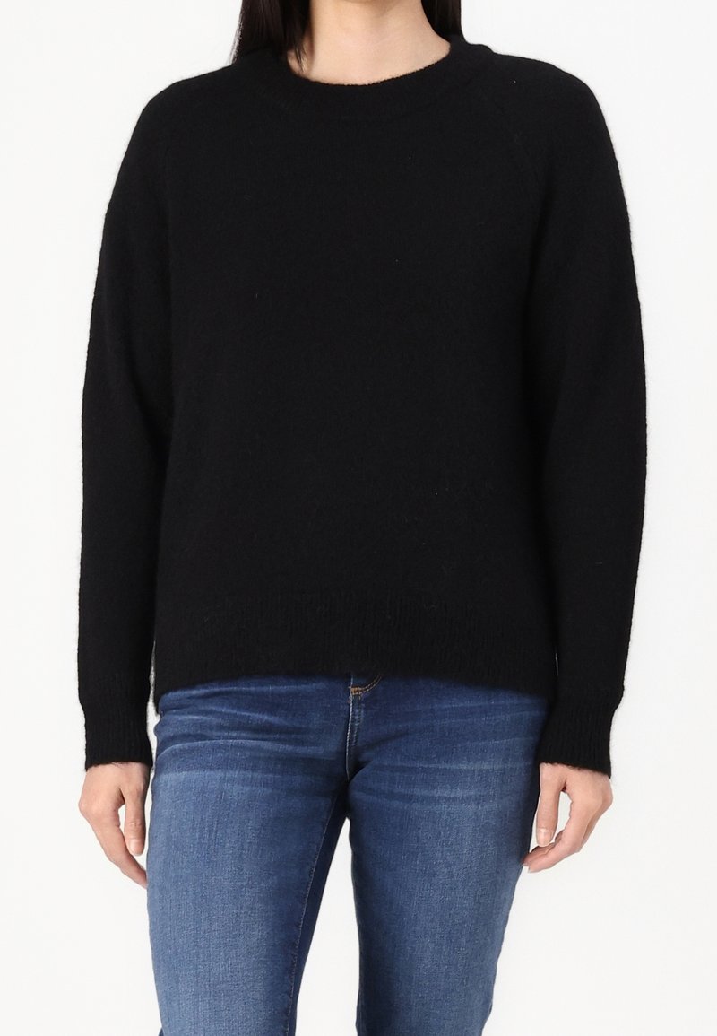 Person in einem schlichten schwarzen Langarmpullover und blauen Jeans, stehend vor einem weißen Hintergrund.