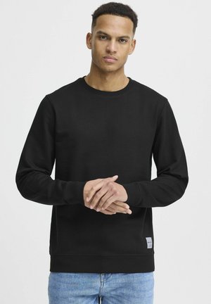 Black crewneck sweatshirt made from cotton, featuring ribbed cuffs and hem, with a small logo tag at the bottom left. Textured, casual design.