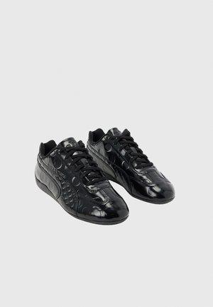 Een paar glanzende zwarte sneakers met geperste cirkelpatronen en zwarte veters, getoond vanuit een voor-zij-aanzicht op een grijze achtergrond.