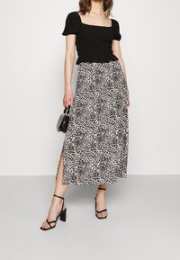 Top smock nero con maniche a sbuffo corte, abbinato a una maxi gonna beige con stampa leopardata e spacchi laterali, e sandali neri con tacco blocco.