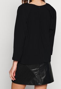 Haut noir à manches longues avec un col froncé, présentant une fermeture éclair discrète dans le dos. Assorti à une jupe en cuir noire mettant en valeur des panneaux diagonaux.