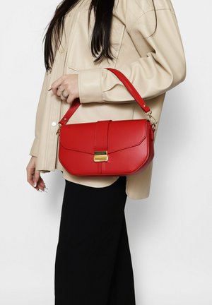 Frau in beigefarbener Jacke, die eine rote Leder-Umhängetasche mit goldenem Verschluss hält, kombiniert mit schwarzer Hose vor weißem Hintergrund.