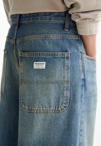 Gros plan sur la poche arrière d’un jean en denim bleu avec une étiquette blanche Pull&Bear, porté avec une chemise beige à manches longues.