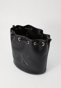 Schwarze Leder-Eimer-Tasche mit geprägtem 'CK'-Logo, goldfarbenen Beschlägen und Zugverschluss. Texturierte Oberfläche mit abgerundetem oberen Rand.