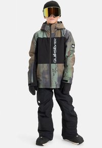 Black and camo snow jacket with logo, paired with black snow pants and gloves. Accessories include tinted goggles and a black beanie.