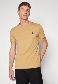 Kortærmet T-shirt med rund hals i en sennepsgul farve, der har et lille sort trælogo på venstre bryst, lavet af et blødt stof.