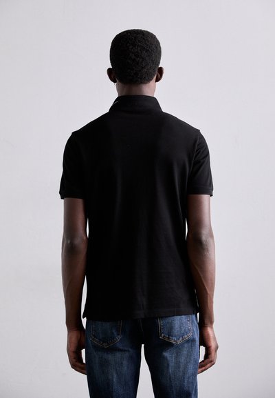 Svart polo med klassisk krage, korta ärmar och en slät textur, tillsammans med blå denimjeans. Bakifrån visas.
