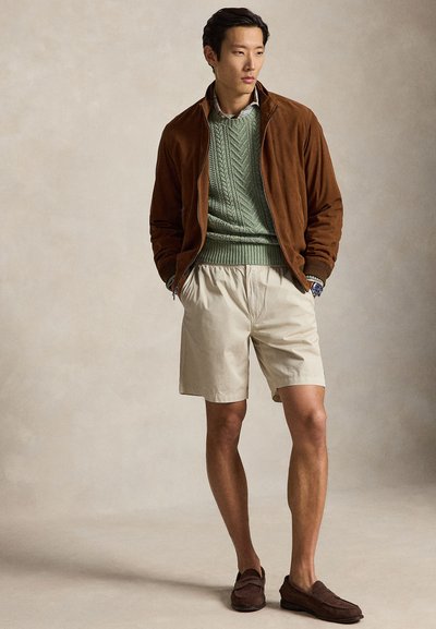 Moški, oblečen v rjavo semiš jakno, zelen pulover s pletenim vzorcem, bež kratke hlače in rjave mokasine, stoji z rokami v žepih.