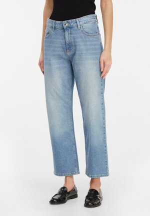 Woman wearing light blue straight-leg jeans, black sleeveless top, and shiny black loafers, standing against a white background.