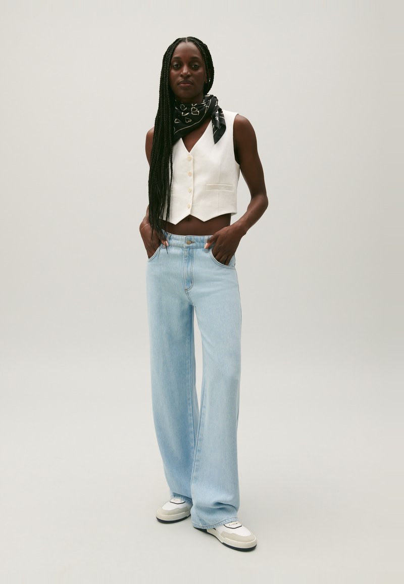 Débardeur blanc court avec un col en V et une fermeture à boutons, associé à un jean large bleu clair et un foulard noir à motifs, porté avec des baskets.