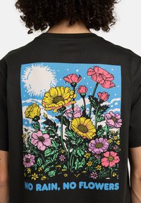 Sort bomulds t-shirt med et farverigt blomsterdesign på ryggen, med gule, lyserøde og lilla blomster, en blå himmel og teksten "INGEN REGN, INGEN BLOMSTER."