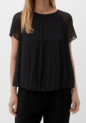 Woman wearing a black pleated chiffon blouse with sheer short sleeves and a round neckline, paired with black pants.
