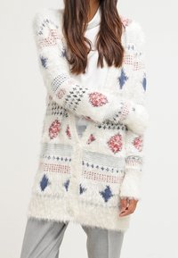 Chlupatý bílý cardigan s barevnými vzory v červené, modré a šedé. Má výstřih do V, zapínání na knoflíky a dlouhé rukávy. Měkká textura.