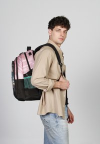 Mochila con secciones en rosa, negro y turquesa; cuenta con un cierre, un bolsillo de malla y correas acolchadas. Llevada en un hombro por una persona.