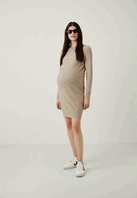 Rochie de maternitate tricotată, bej, cu siluetă ajustată, mâneci lungi, până la genunchi, asortată cu adidași albi. Textură netedă și design simplu.