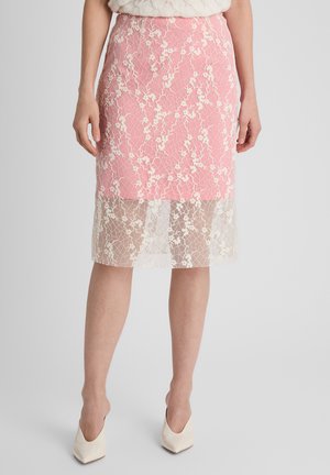 Vrouw draagt een knielange roze rok met een witte bloemen kanten overlay en witte puntige hakken, staand tegen een effen achtergrond.