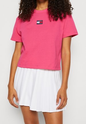 Kobieta ubrana w różowy krótki t-shirt Tommy Jeans i białą plisowaną spódnicę, stoi z ramionami swobodnie opuszczonymi wzdłuż ciała.