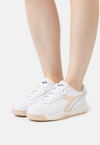 White athletic sneakers with beige accents, a perforated texture, flat laces, and a padded collar. Features a rubber sole and curved design.