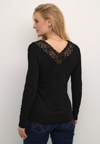 Maglietta nera a maniche lunghe con scollatura a V e dettagli in pizzo sulla schiena. Tessuto morbido con un design aderente. Accentata da bordi ondulati.
