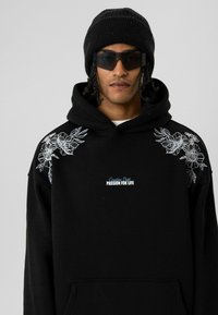 Zwarte hoodie met bloemrijke borduring op de schouders, voorzien van een voorzak en tekst "Creative Dept. Passion for Life" in blauw. Gecombineerd met een zwarte muts en zonnebril.