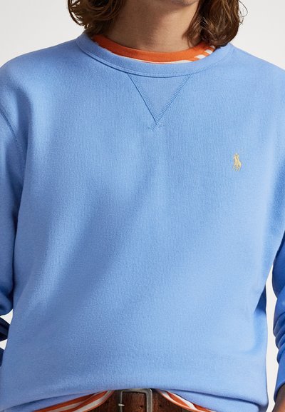 Ανοιχτό μπλε φούτερ με στρογγυλή λαιμόκοψη και μανίκια με μανσέτες, με μικρό κίτρινο λογότυπο στο στήθος και φορεμένο πάνω από μια πορτοκαλί ριγέ πουκάμισο.