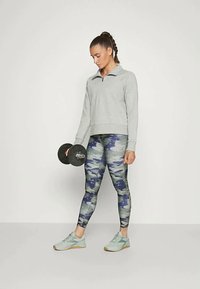 Pelēks sporta kapučs, rakstainas legingi zilā un pelēkā krāsā, gaiši zilas sporta apavi un melna hanteles rokā.