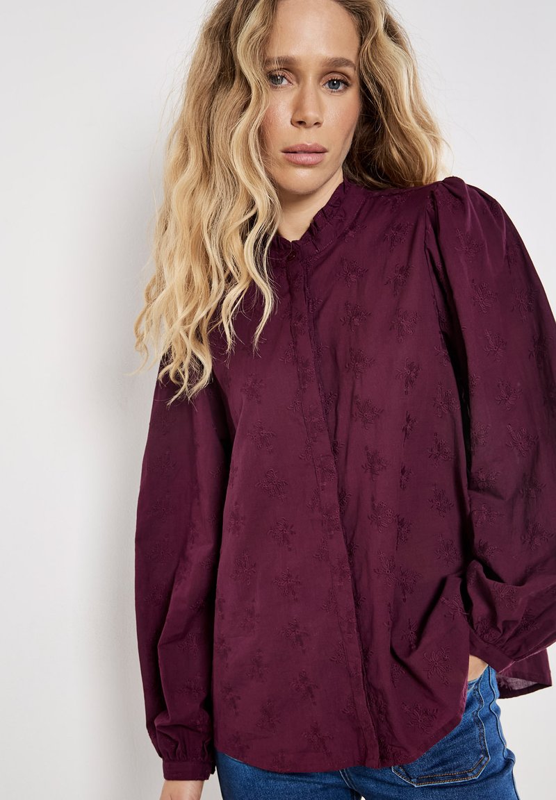 Blusa burdeos con mangas largas abullonadas, tejido texturizado con bordado floral y cuello redondo cerrado. Combina con unos jeans azul.