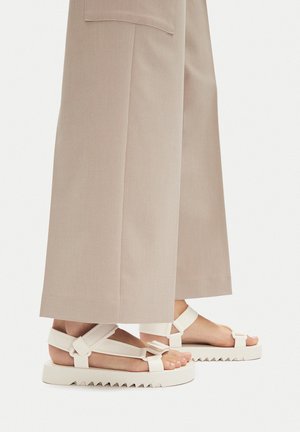 Białe sandały z masywną podeszwą o zygzakowatych krawędziach, szerokimi paskami na stopie oraz małą kokardką na pasku wokół kostki.