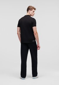 Polo nero con maniche corte e orlo dritto, abbinato a pantaloni neri, mostrato da dietro su uno sfondo grigio chiaro.