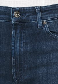 Vaqueros de mezclilla azul oscuro con cintura alta, presentando un cierre de botón plateado, costura decorativa y un bolsillo frontal. Textura suave.