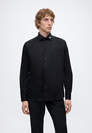 Junger Mann mit lockigem Haar, der ein schwarzes Hemd mit Knöpfen und einem kleinen weißen Logo am Kragen trägt, steht vor einem schlichten weißen Hintergrund.