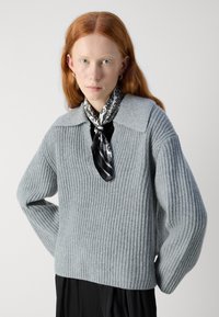 Világosszürke kötött pulóver, bordázott textúrával, széles ujjakkal és galléros nyakkal. Fekete-fehér mintás sál van megkötve a nyakán.