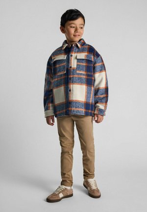 Veste à carreaux en bleu, crème et orange ; texture en polaire avec deux poches poitrine. Un pantalon beige et des baskets marron complètent le look.