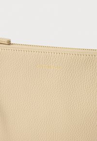 Bež kožna torbica s teksturiranom površinom i zlatnim utisnutim logotipom. Ima zatvarač na vrhu i glatku tkaninu na unutrašnjosti.