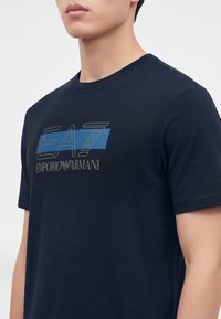 Άνδρας που φοράει μπλε σκούρο κοντομάνικο μπλουζάκι με το λογότυπο "EA7 Emporio Armani" σε μπλε και γκρι χρώμα στο στήθος, σε μονόχρωμο φόντο.
