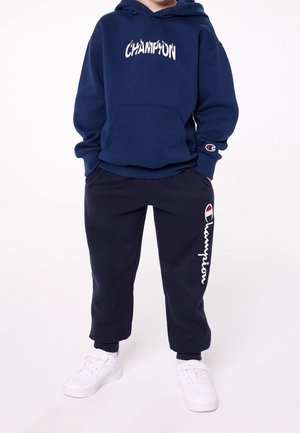 Kind draagt een marineblauwe Champion hoodie en trainingsbroek met witte logo’s, handen in de zakken en witte sneakers, staand op een witte achtergrond.
