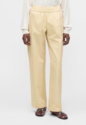 Personne portant un pantalon beige droit avec une taille élastique et un haut blanc à manches longues, debout avec des sandales noires aux pieds.