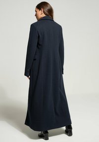 Donna di spalle che indossa un lungo cappotto blu navy con tasche laterali, stivali neri con tacco e orecchini a cerchio dorati, su uno sfondo grigio semplice.