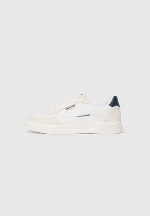 Hvid lav Sneaker med beige ruskind accents, sort hæl fanen, hvide snørebånd og "just cavalli" label på siden mod en lysegrå baggrund.