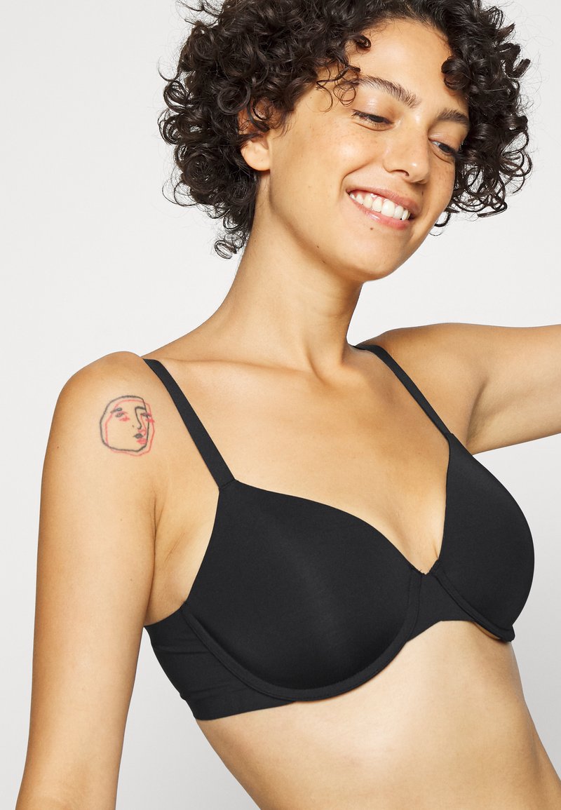 Schwarzer gepolsterter BH mit verstellbaren Trägern, der ein glattes Design und einen V-Ausschnitt aufweist. Sichtbares Tattoo auf der Schulter.