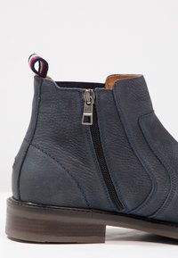 Navy kožené kotníkové boty s texturovaným povrchem, bočním zipem a kontrastním poutkem. Mají robustní hnědou koženou podrážku a šité detaily.