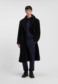 Abrigo negro de doble botonadura sobre una sudadera azul marino y pantalones a juego. El abrigo tiene una textura suave y solapas acanaladas, combinado con zapatillas negras.