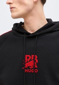 Κοντινό πλάνο ενός ατόμου που φοράει μαύρο hoodie με το λογότυπο της Red Bull και το κείμενο «HUGO» τυπωμένα στο στήθος.