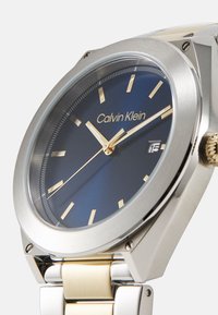 Reloj Calvin Klein con esfera azul, marcadores de hora en tono dorado, caja de acero inoxidable plateado y pulsera de metal de dos tonos con ventana de fecha.