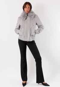 Donna con un giubbotto grigio chiaro con colletto di pelliccia, pantaloni neri a zampa d'elefante e décolleté neri a punta, in piedi su uno sfondo bianco.