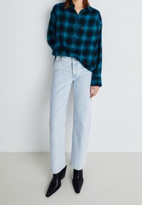 Camicia a quadri teal e nera con bottoni, abbinata a jeans dritti azzurri chiari e stivali neri con tacco a punta. Branding sottile visibile.