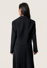 Czarna, dopasowana marynarka o wydłużonej sylwetce, z usztywnionymi ramionami i zapinanymi mankietami, w zestawie z dopasowaną długą sukienką. Gładka tekstura materiału.
